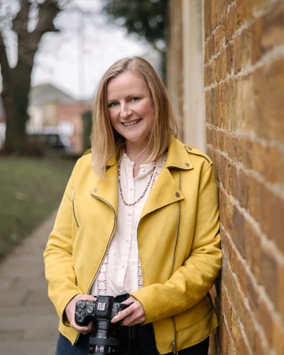 Claire in a yellow jacket holding her camera beside a brick wall