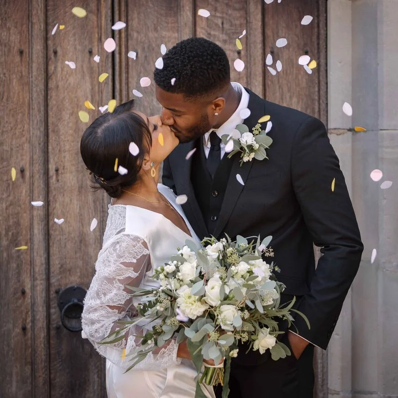 A wedding couple kissing beside a wooden door as confetti falls