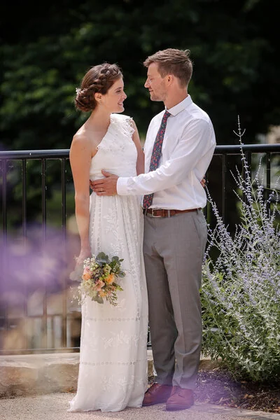 A wedding couple holding hands beside purple summer flowers