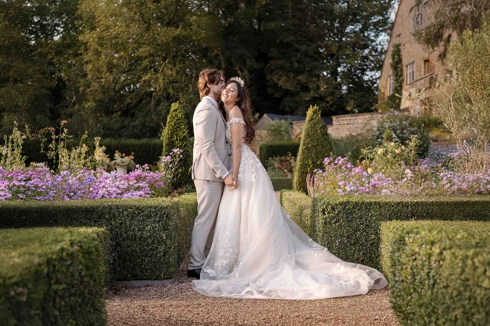 A wedding couple embracing beside clipped hedges and purple garden flowers