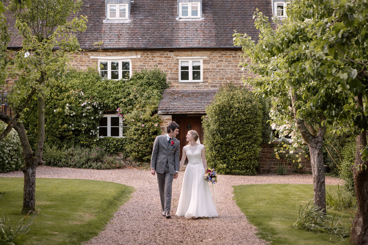 Bride and groom walking together in front of Dodmoor House, Northamptonshire, on their wedding day.