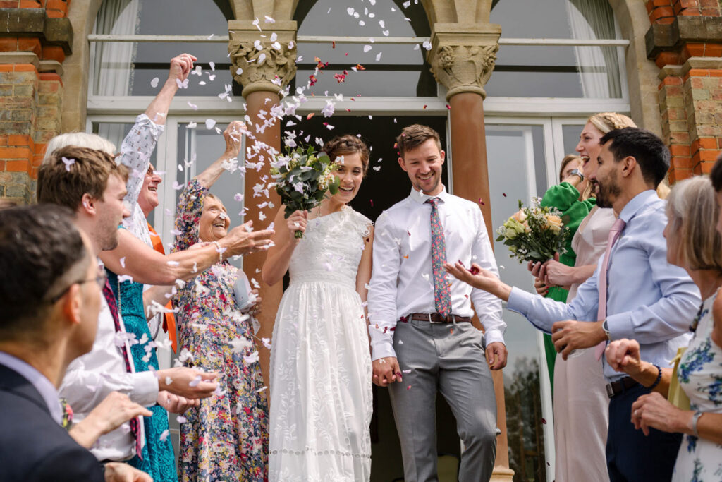 A bride and groom looking happy on their wedding day as they walk down the steps through a flurry of confetti at St. Hilda's College, Oxford.