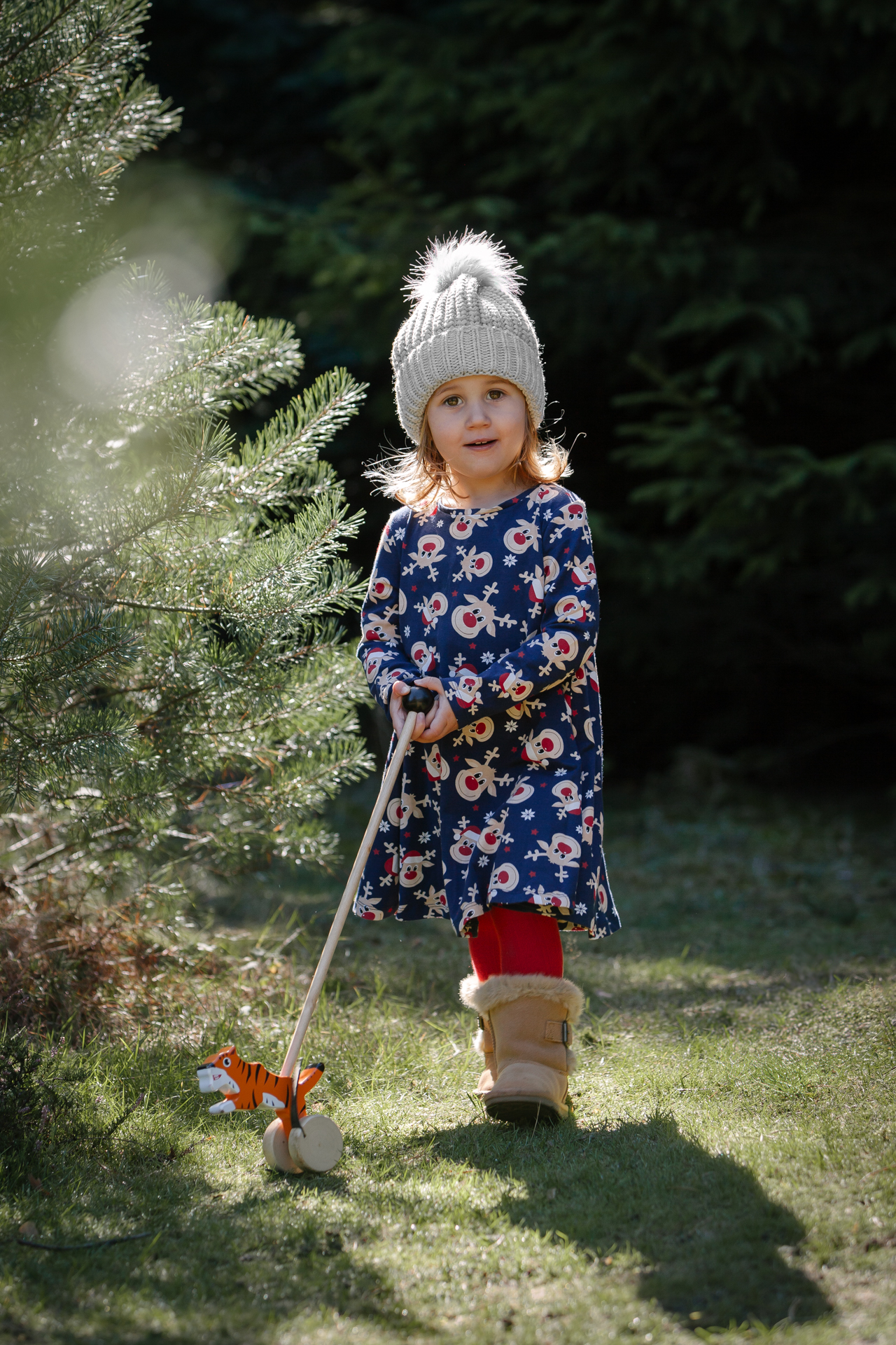 Child pushing a wooden toy in a Christmas tree field during an outdoor mini photoshoot, wearing a woolly hat, Christmas dress, and boots.