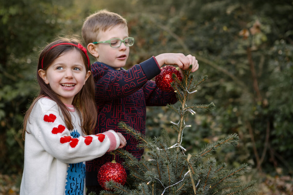 Boy and girl decorating a Christmas tree during an outdoor Christmas mini-shoot in Northamptonshire, with the girl smiling at the camera.