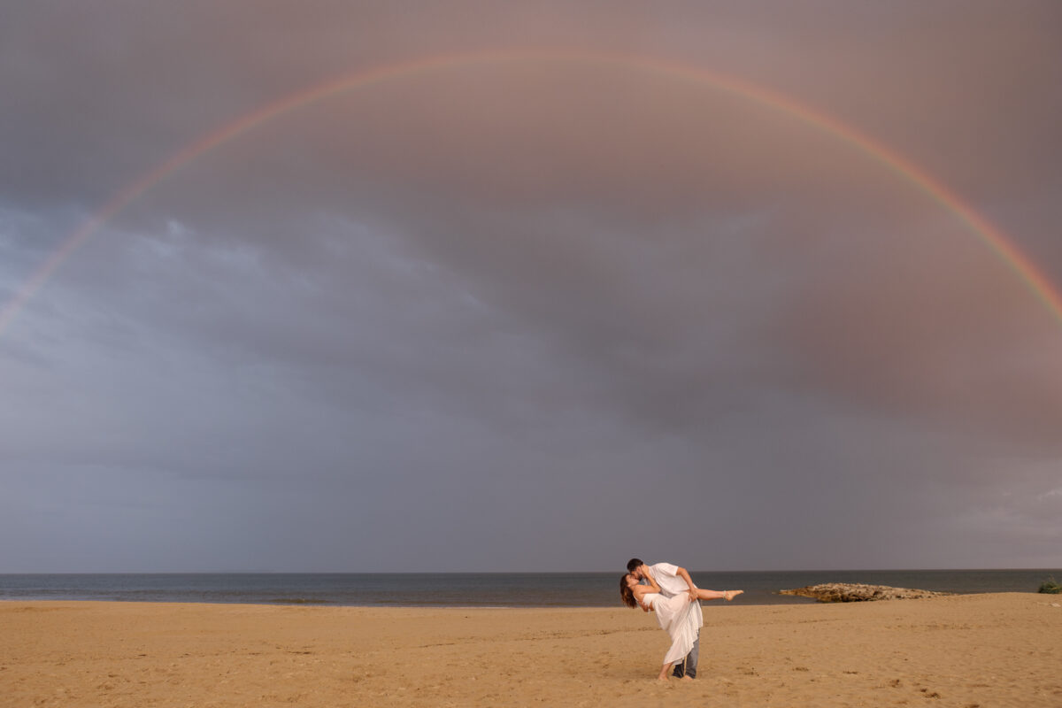 Couple dip-kissing under a rainbow on Sandbanks Beach, Dorset, wearing ideal attire for a beach engagement photoshoot, including a flowing dress and casual shirt.